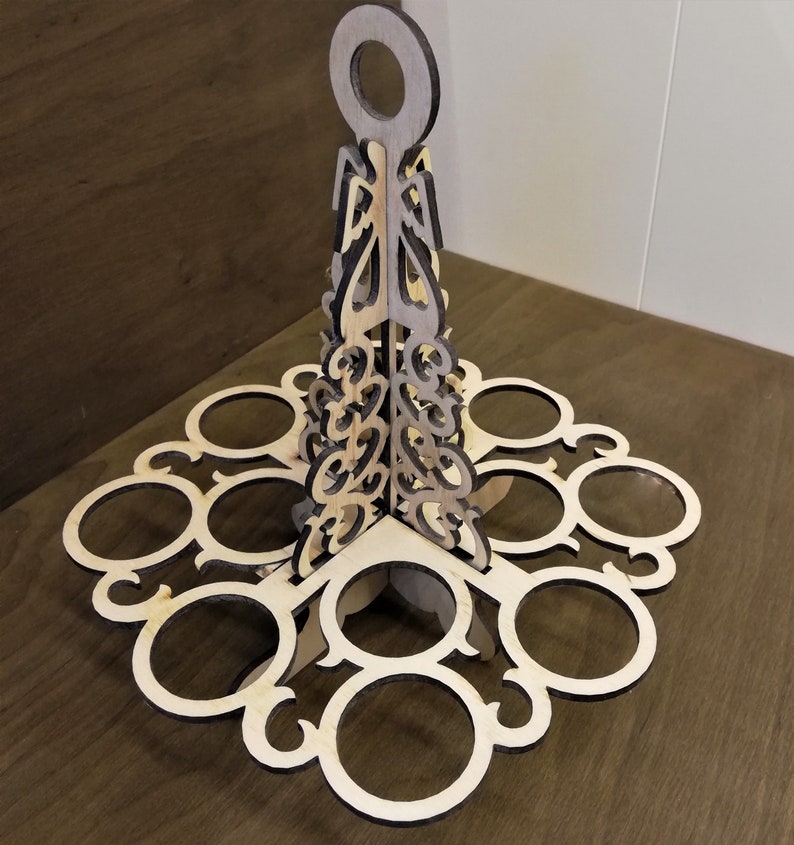 May include: A wooden egg holder with a decorative design. It has a handle at the top and eight slots for eggs. The holder is made of light brown wood with a darker brown stain.