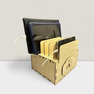 Könnte beinhalten: Eine hölzerne Ladestation mit mehreren Schlitzen für Tablets und Smartphones. Geräte sind eingesetzt und mit Ladekabeln verbunden. Die Station hat ein wolkenförmiges Logo mit dem Text "LET'S RECHARGE".