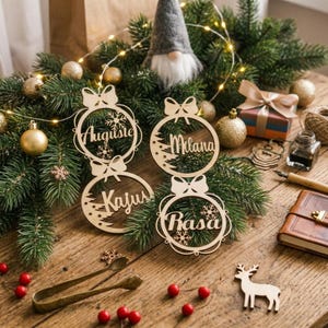 May include: Four circular wooden Christmas ornaments featuring cut-out designs of bows, snowflakes, and pine trees. Each ornament is laser-cut with a unique name: Auguste, Milana, Kajus, and Rasa. The ornaments are arranged on a wooden surface.