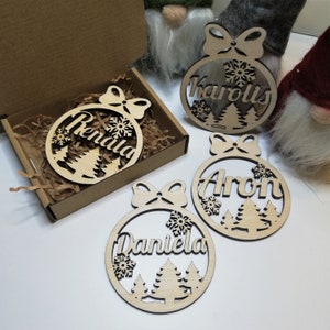 May include: Four wooden Christmas ornaments with a winter scene design. Each ornament features a different name: "Rendala", "Karolis", "Aron", and "Daniela".
