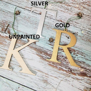 May include: Three wooden letters, K, T, and R, are shown on a light blue wood background. The letter K is unpainted, the letter T is painted silver, and the letter R is painted gold. Each letter has a hole at the top for hanging.