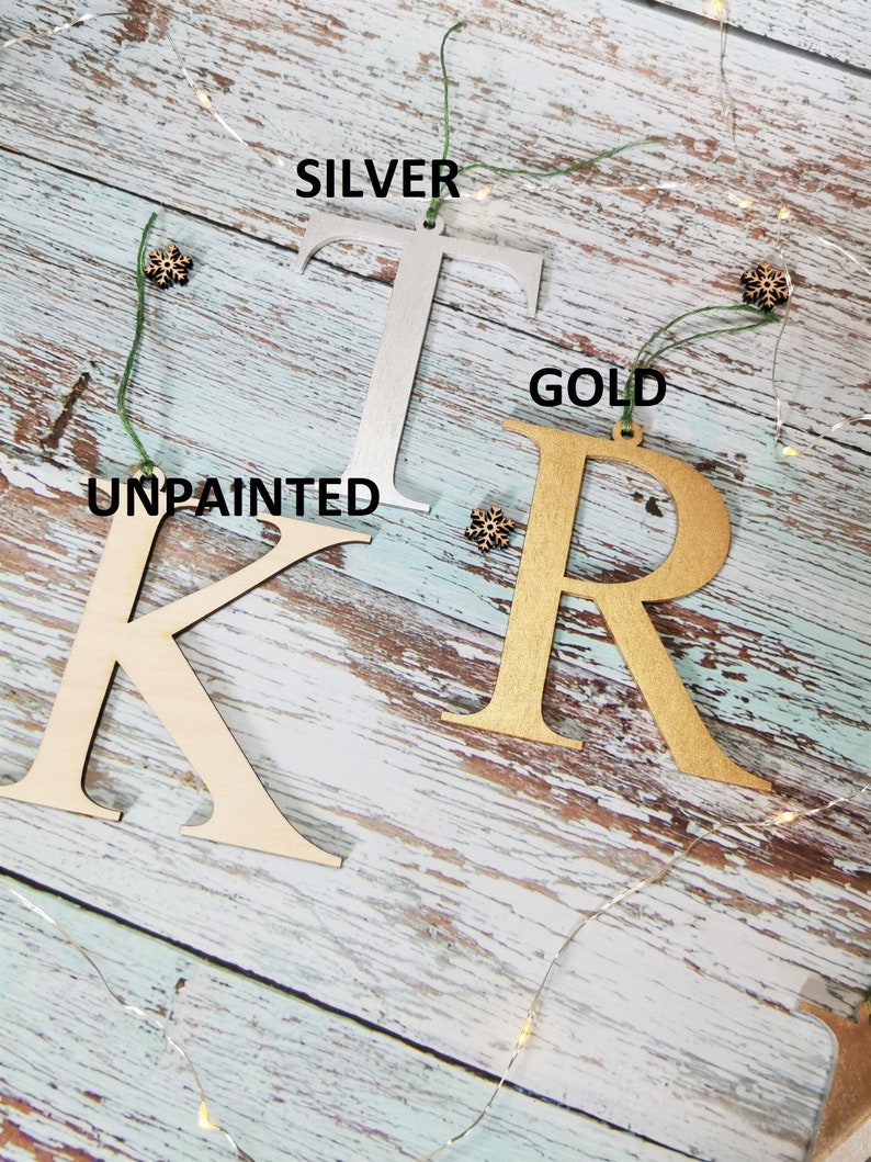 May include: Three wooden letters, K, T, and R, are shown on a light blue wood background. The letter K is unpainted, the letter T is painted silver, and the letter R is painted gold. Each letter has a hole at the top for hanging.