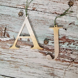 May include: Two wooden Christmas decorations in the shape of the letters A and J. The letters are painted gold and white and have a rustic look. The decorations are hanging from twine.
