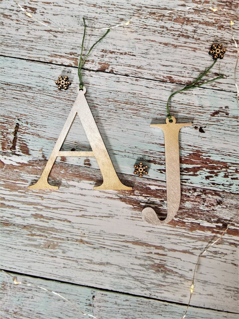 May include: Two wooden Christmas decorations in the shape of the letters A and J. The letters are painted gold and white and have a rustic look. The decorations are hanging from twine.