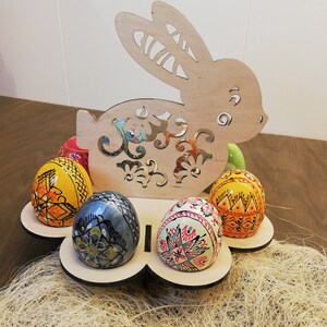 May include: Wooden bunny shaped Easter egg holder with six painted eggs. The eggs are decorated with colorful geometric patterns and designs. The bunny is cut out and has a floral design.
