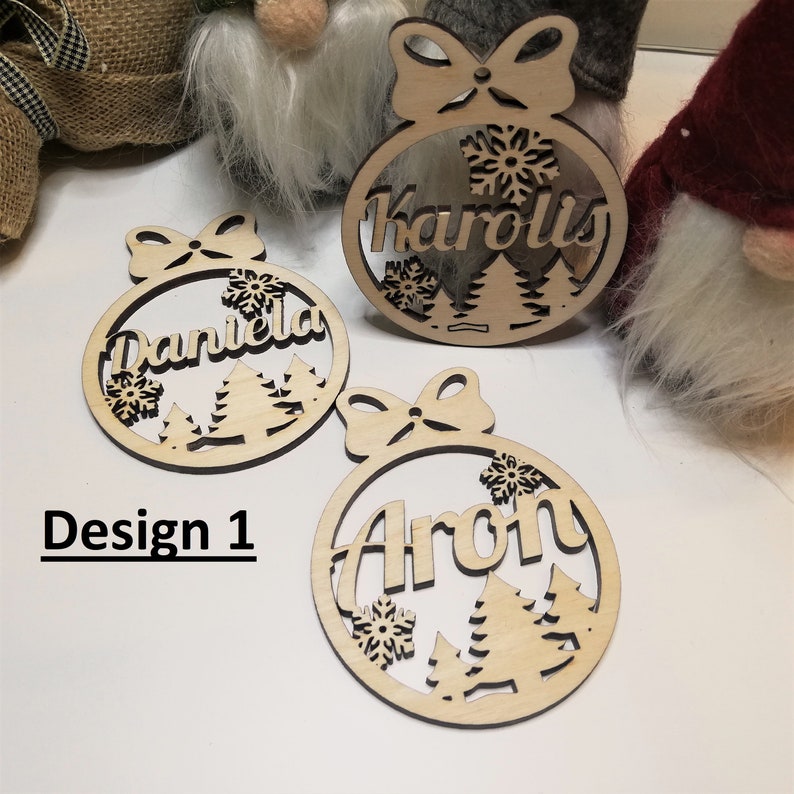 May include: Three wooden Christmas ornaments with a snowflake and tree design. The ornaments are personalized with the names "Karolis", "Daniela", and "Aron".