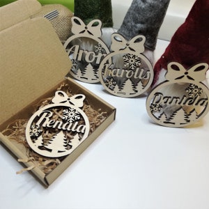 May include: Three wooden Christmas ornaments with names "Aron", "Karolis", and "Daniela" written on them. The ornaments are decorated with snowflakes and pine trees. One ornament is in a brown box with shredded paper.