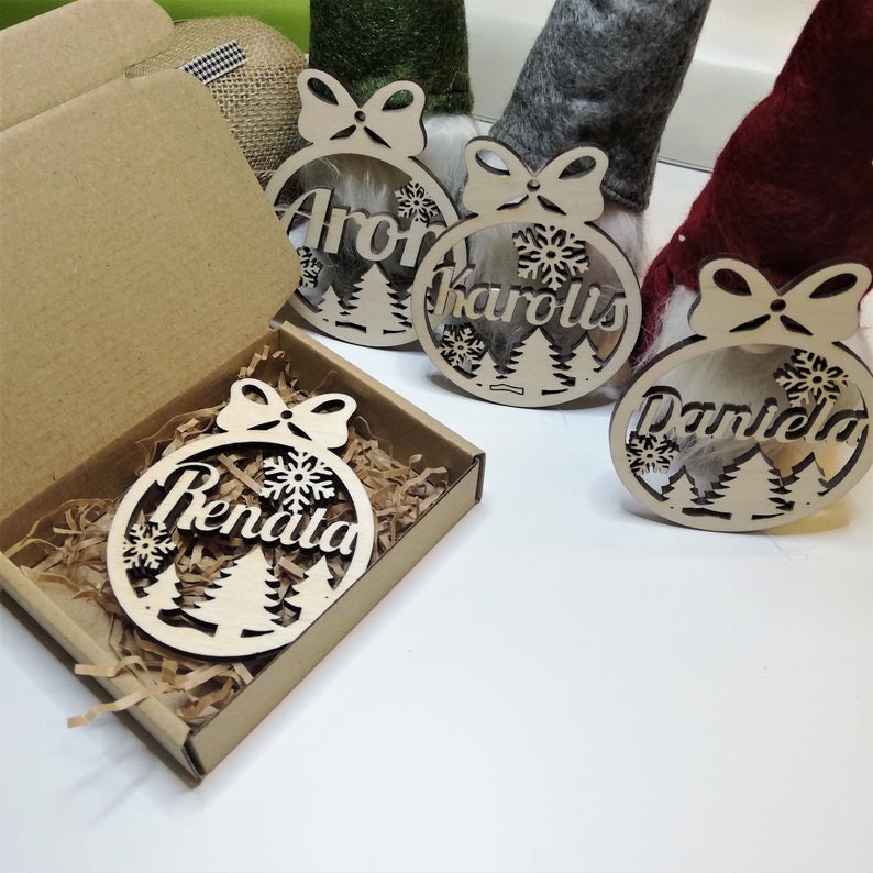May include: Three wooden Christmas ornaments with names "Aron", "Karolis", and "Daniela" written on them. The ornaments are decorated with snowflakes and pine trees. One ornament is in a brown box with shredded paper.