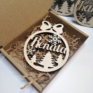 May include: A wooden Christmas ornament shaped like a circle with a bow at the top. The ornament has a cutout design of a Christmas tree and snowflakes. The name "Renata" is written in the center of the ornament.