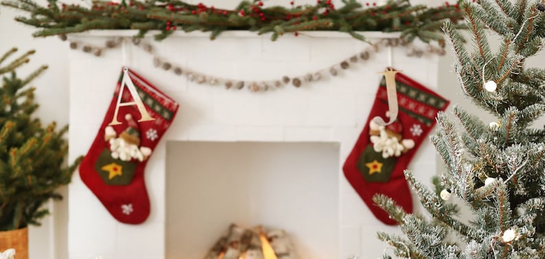 May include: Two red Christmas stockings with white trim and a snowman design hang from a white fireplace mantel. The stockings are decorated with gold stars and snowflakes. A garland of white beads hangs above the stockings.