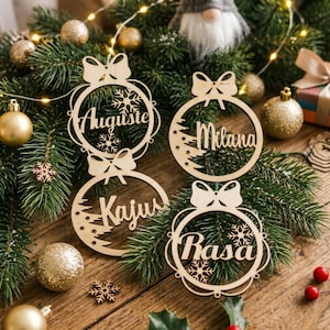 May include: Four circular light-colored wooden ornaments featuring cut-out designs of bows, snowflakes, and pine trees. Each ornament is personalized with a name in cursive script: Auguste, Milana, Kajus, and Rasa, arranged on green pine branches.