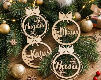 May include: Four circular light-colored wooden ornaments featuring cut-out designs of bows, snowflakes, and pine trees. Each ornament is personalized with a name in cursive script: Auguste, Milana, Kajus, and Rasa, arranged on green pine branches.