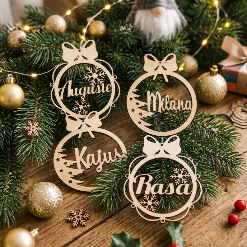 May include: Four circular light-colored wooden ornaments featuring cut-out designs of bows, snowflakes, and pine trees. Each ornament is personalized with a name in cursive script: Auguste, Milana, Kajus, and Rasa, arranged on green pine branches.