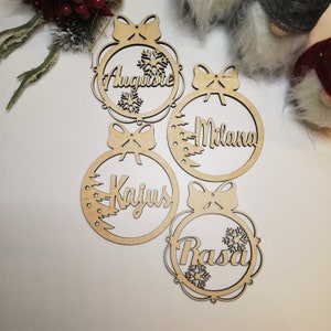 May include: Four wooden Christmas ornaments with names "Augustine", "Milana", "Kajus", and "Rasa" written on them. Each ornament has a bow and a snowflake design.
