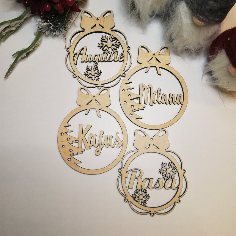 May include: Four wooden Christmas ornaments with names "Augustine", "Milana", "Kajus", and "Rasa" written on them. Each ornament has a bow and a snowflake design.