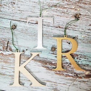 May include: Three wooden letters, K, R, and T, painted in gold and silver, are strung with green twine and hung on a rustic wooden background. The letters are Christmas ornaments.