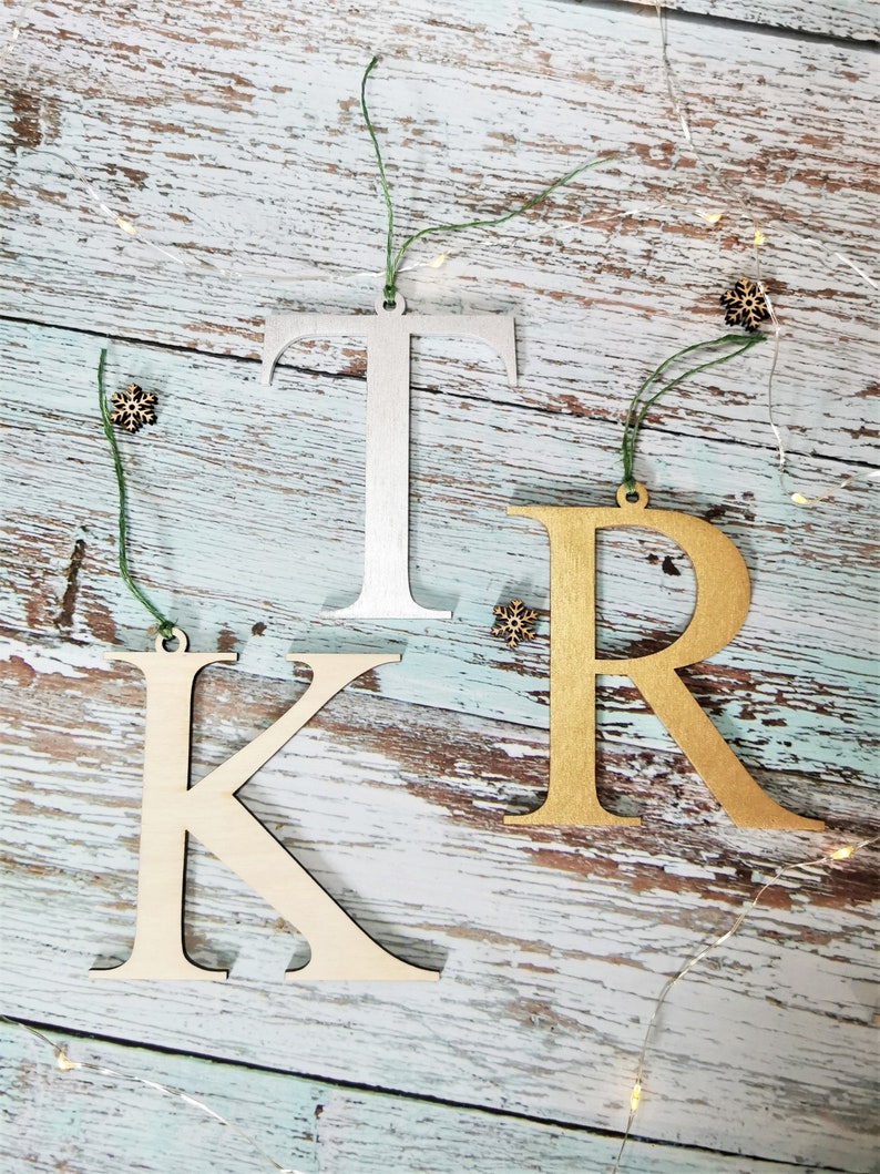May include: Three wooden letters, K, R, and T, painted in gold and silver, are strung with green twine and hung on a rustic wooden background. The letters are Christmas ornaments.