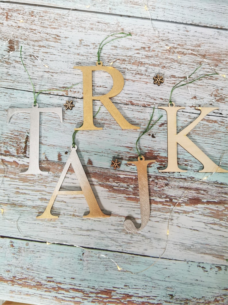 May include: Wooden letters spelling out the name "TAJ" in gold and silver paint, with the letter "R" and "K" in gold paint. The letters are hanging from twine on a rustic wooden background.
