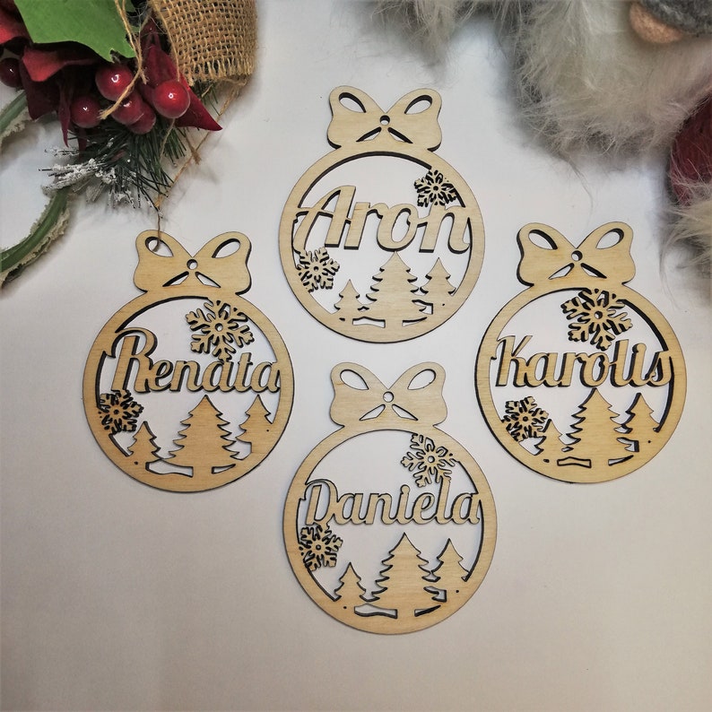 May include: Four wooden Christmas ornaments with names "Aron", "Renata", "Karolis", and "Daniela" written on them. Each ornament features a bow, snowflakes, and pine trees.