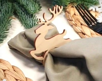 Puede incluir: Un servilletero de madera con forma de reno adorna una servilleta gris doblada. El reno es de color marrón claro y tiene cuernos detallados. Un salvamanteles tejido y ramas de hoja perenne sugieren un tema navideño.