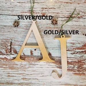 May include: Two wooden letters, A and J, painted in gold and silver. The letters are hanging from twine and have a rustic wood background.