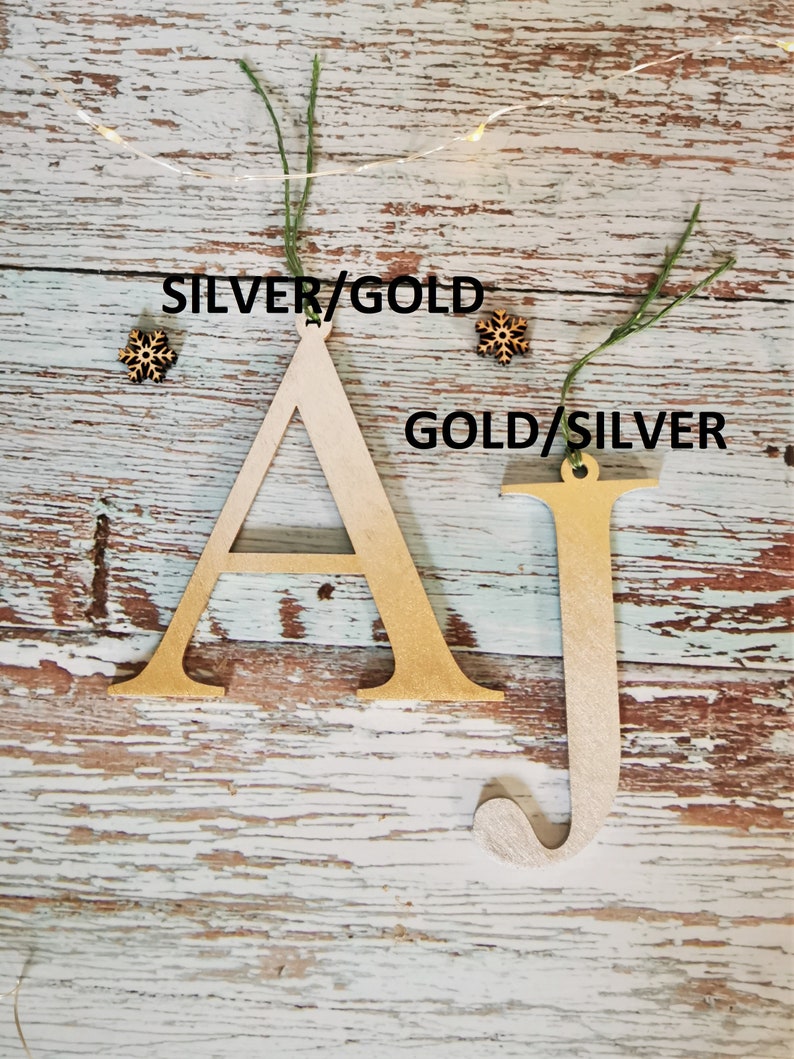 May include: Two wooden letters, A and J, painted in gold and silver. The letters are hanging from twine and have a rustic wood background.