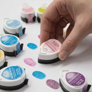May include: A hand presses a pink ink pad onto white paper, creating a fingerprint. Several other ink pads in various colors, including blue, purple, and yellow, are scattered around, along with their corresponding fingerprints. The ink pads are labeled "Memento".
