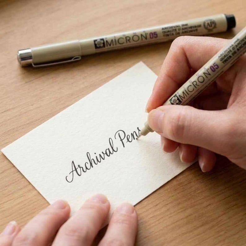 Puede incluir: Una mano escribe "Archival Pens" en una tarjeta color crema con un bol&iacute;grafo de tinta de archivo Micron 05. Otro bol&iacute;grafo est&aacute; en el fondo. Los bol&iacute;grafos son beige con tinta negra y detalles plateados. La tarjeta est&aacute; sobre una superficie de madera.