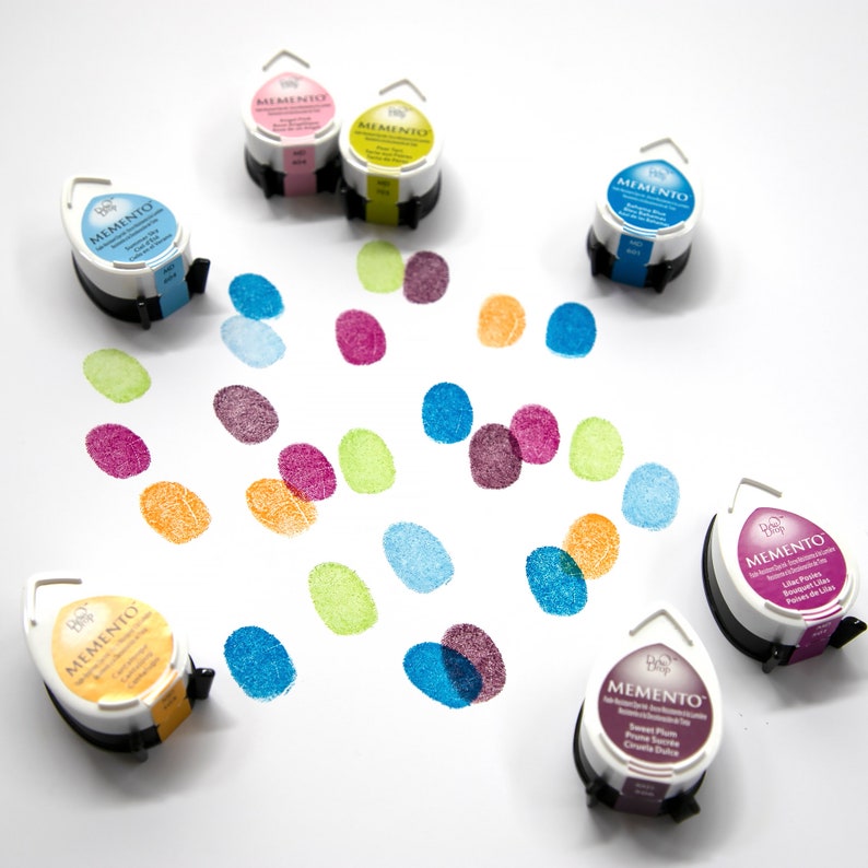 May include: A collection of Memento ink pads in various colors, including blue, pink, yellow, and purple, with corresponding fingerprint impressions. The ink pads are white with black bases and feature the Memento logo.