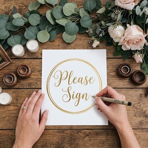 Puede incluir: Escena de boda con una mesa de madera, con un letrero que dice "Please Sign" en letras doradas. La escena incluye velas, un marco de fotos, anillos y flores, creando un ambiente rom&aacute;ntico.