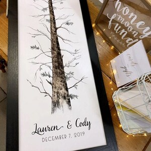 May include: A black framed print of a tree with the names Lauren & Cody and the date December 7, 2019. The print is on a wooden surface with a white box that says "cards and gifts" and a wooden box that says "honeymoon fund".