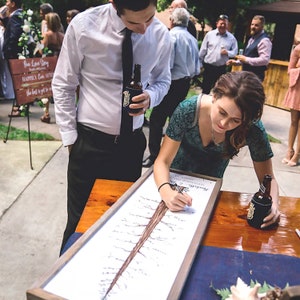 May include: A couple signs a tree-shaped guest book at a wedding reception. The guest book is on a wooden table with a blue tablecloth. The couple is standing next to the table, and the groom is holding a bottle of beer.