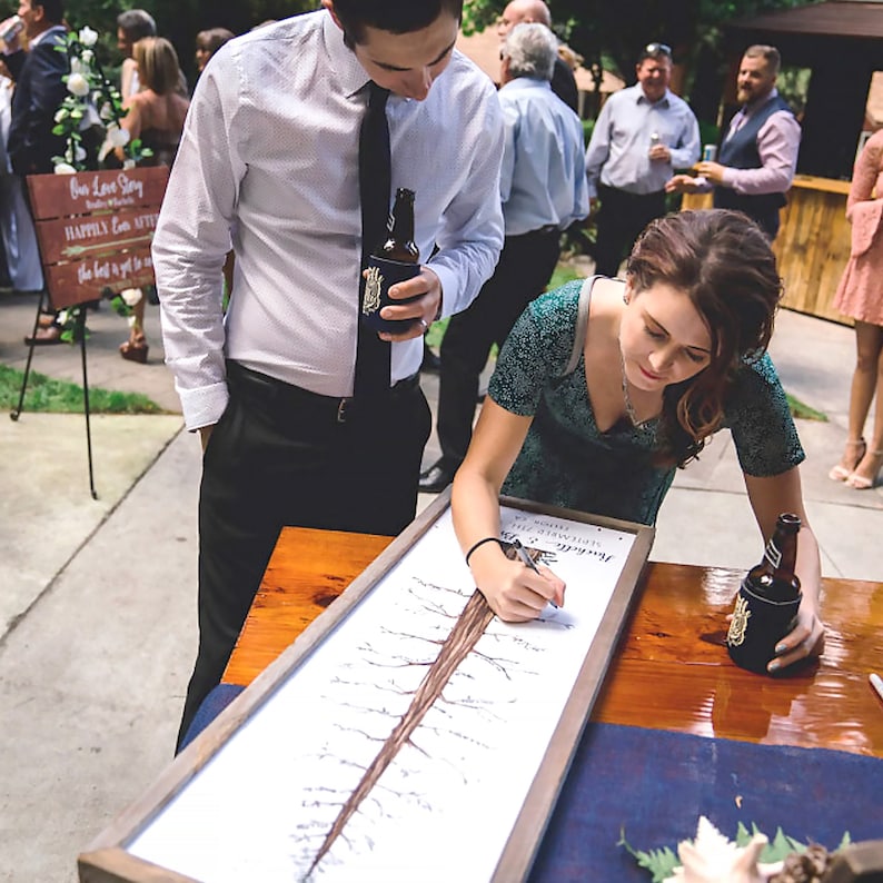 May include: A couple signs a tree-shaped guest book at a wedding reception. The guest book is on a wooden table with a blue tablecloth. The couple is standing next to the table, and the groom is holding a bottle of beer.