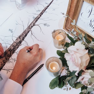 May include: A person signing a tree drawing on a white table with a sign that says "Please Sign" in calligraphy. There are two candles and a bouquet of pink roses on the table.