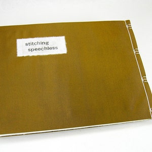 Puede incluir: Un libro de tapa dura marrón con costuras blancas a lo largo del lomo. La portada tiene una etiqueta blanca con el texto "stitching speechless" impreso en negro.