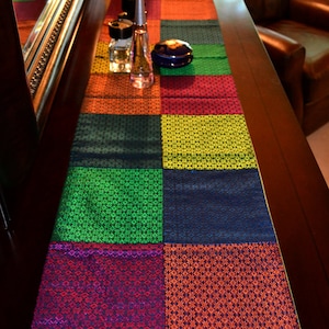 May include: A colorful table runner with a pink border and a patchwork design of green, blue, yellow, orange, and red squares. The runner is made of woven fabric with a subtle pattern.