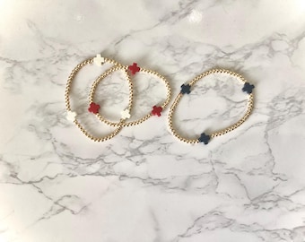 Puede incluir: Tres pulseras elásticas hechas de pequeñas cuentas de tono dorado. Dos pulseras presentan dijes en forma de cruz que alternan entre blanco y rojo. La tercera pulsera tiene dijes en forma de cruz de color azul oscuro espaciados uniformemente.