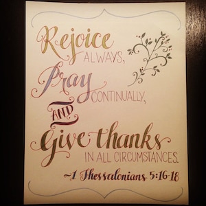 May include: Hand-lettered calligraphy art print with the quote "Rejoice always, pray continually, and give thanks in all circumstances." - 1 Thessalonians 5:16-18. The quote is written in a decorative script with a floral design in the upper right corner.