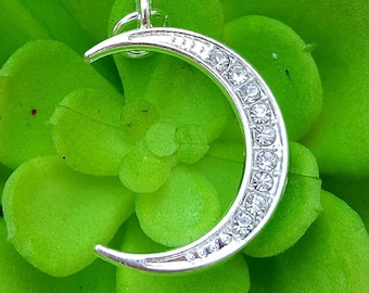 May include: A silver crescent moon charm with clear rhinestones set around the edge. The charm is on a green background.