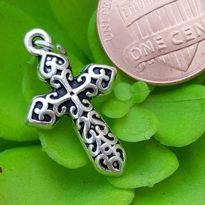 May include: Silver cross charm with an ornate design, next to a US penny for size comparison. The cross is about the size of a penny.