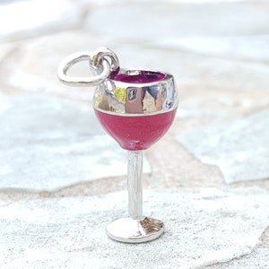 May include: A silver charm in the shape of a wine glass with a red enamel fill. The charm has a loop at the top for attaching to a bracelet or necklace.