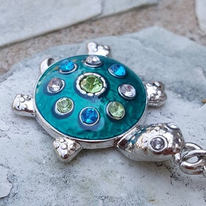 May include: A silver charm in the shape of a turtle with a green enamel shell and blue and green rhinestones. The charm has a silver loop for attaching to a bracelet or necklace.