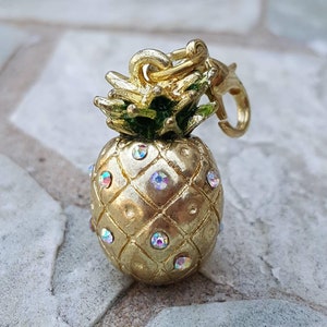 May include: A gold pineapple charm with iridescent rhinestones. The charm has a green top and a gold loop for attaching to a necklace or bracelet.