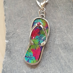 May include: A silver charm in the shape of a flip-flop with a tropical floral print in shades of pink, green, and blue. The flip-flop has a red rhinestone in the center.