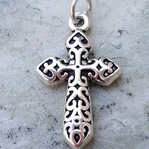 May include: Silver-toned cross charm with an intricate, ornate design. The cross has a loop at the top for attaching to a necklace or bracelet.
