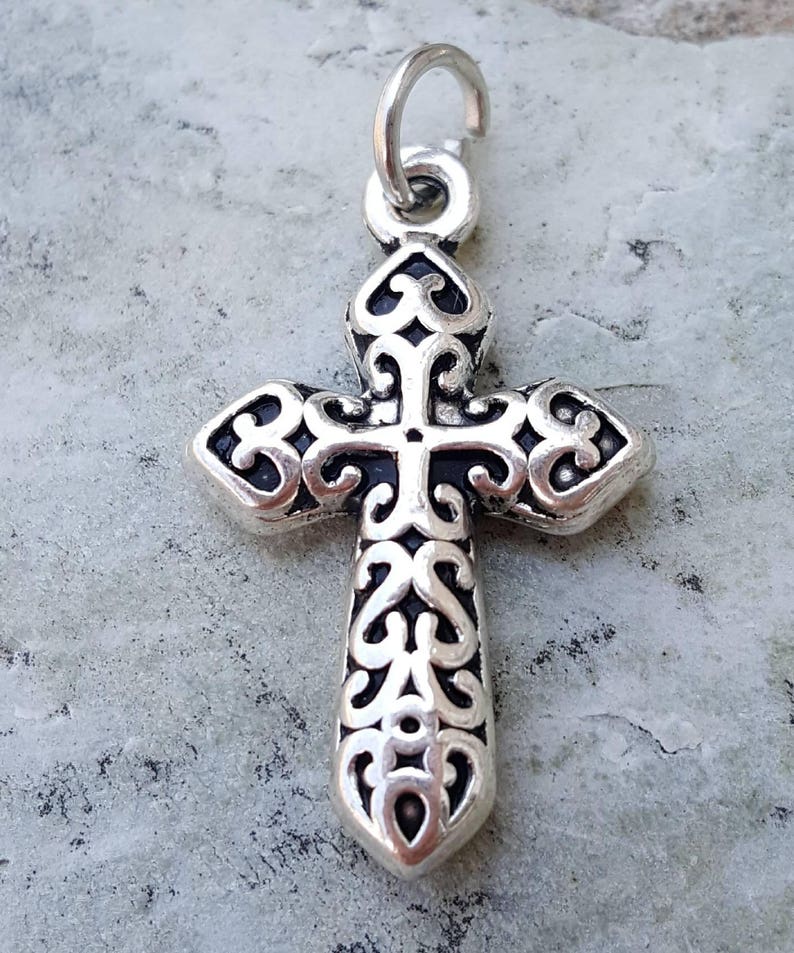 May include: Silver-toned cross charm with an intricate, ornate design. The cross has a loop at the top for attaching to a necklace or bracelet.