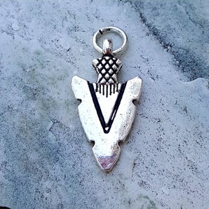 Puede incluir: Encanto de punta de flecha de plata con un diseño decorativo. El encanto tiene un bucle en la parte superior para sujetarlo a un collar o pulsera.