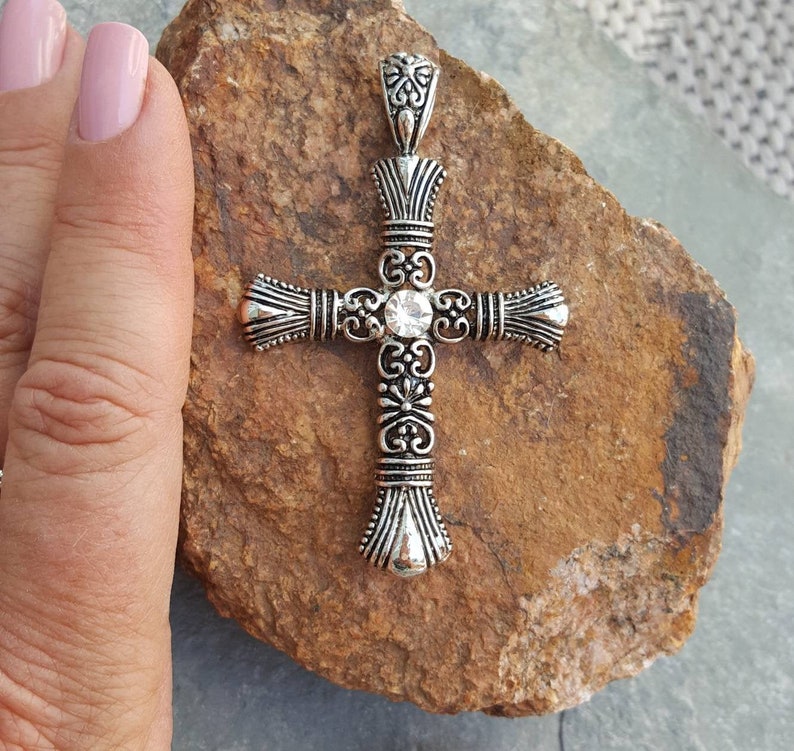 May include: A silver cross pendant with an ornate design and a clear rhinestone in the centre.