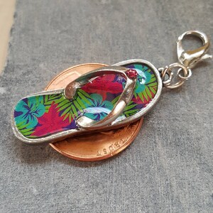 May include: A silver charm in the shape of a flip-flop with a tropical floral print. The charm has a red jewel accent and is attached to a silver split ring.