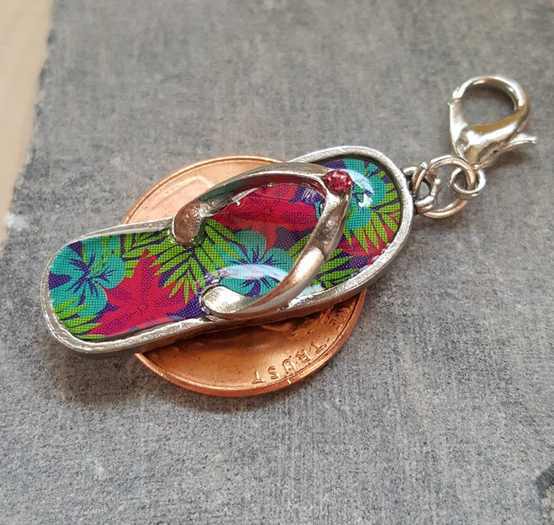 May include: A silver charm in the shape of a flip-flop with a tropical floral print. The charm has a red jewel accent and is attached to a silver split ring.
